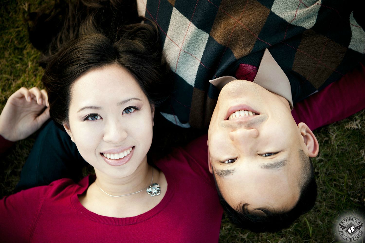 Asian girl with dark hair wearing a red blouse with an infectious smile lies in grass next to a Asian brunette guy  wearing a  checkered sweater at Zilker Park in this flipped engagement photo in Austin.